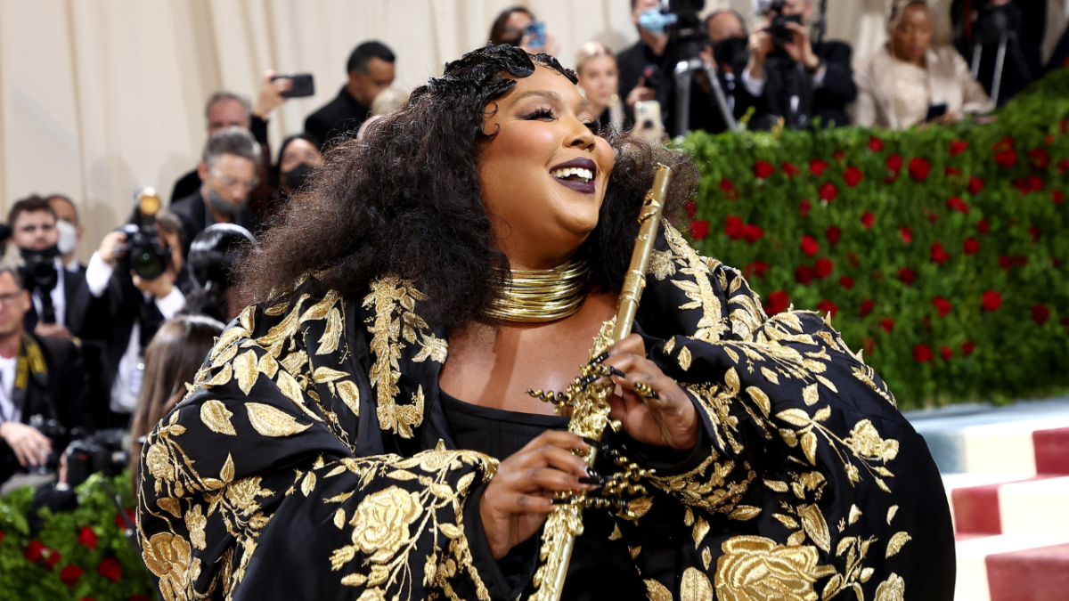 Lizzo at the 2022 Met Gala wearing a black robe with ornate gold flowers and gold jewellery, holding a golden flute.