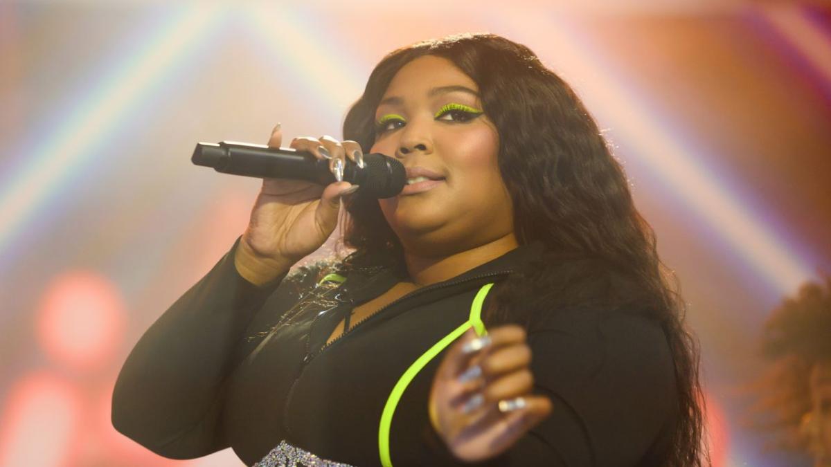 Lizzo holding a microphone with her life hand and smiling into a camera. she is wearing green eyeliner and has green nails.
