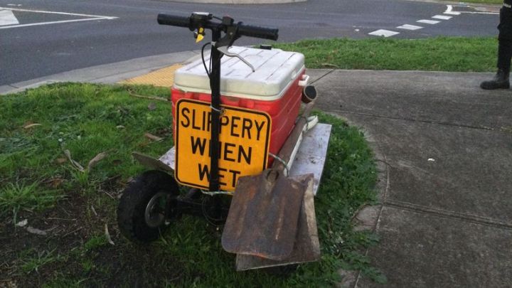 A photo of a red Esky on a scooter with a 'Slippery when wet' warning sign on it and two shovels