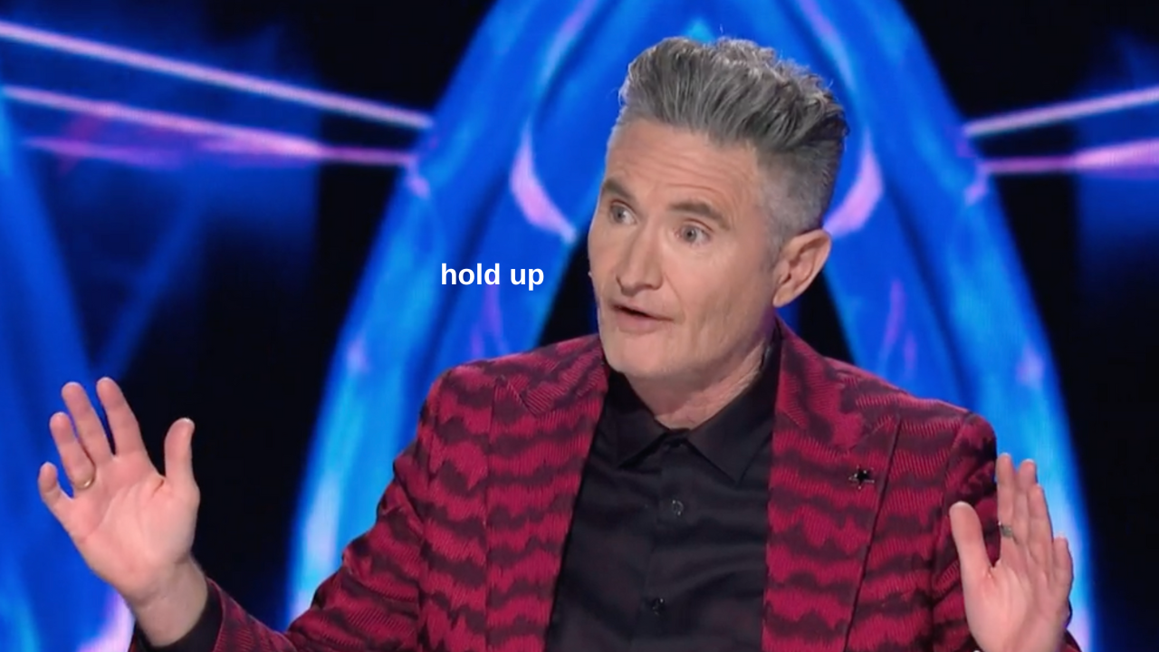 Comedian Dave Hughes on The Masked Singer judging panel wearing a striped red and black jacket and black shirt