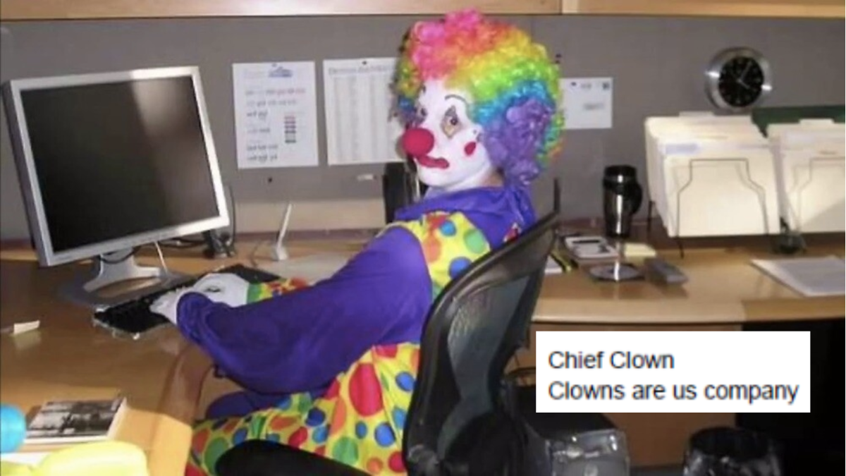 person dressed as clown in purple top and spotted vest, white face makeup, multicoloured wig and red nose seated at computer with screenshot of letter that reads "Chief Clown, Clowns are us company"