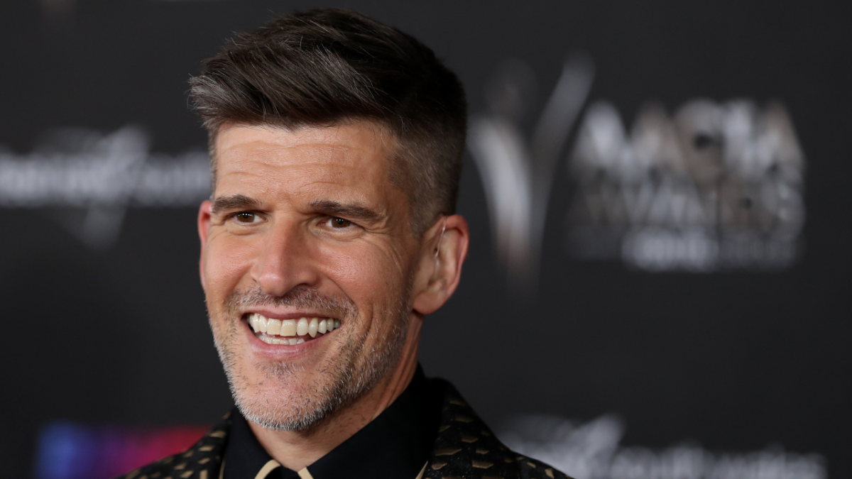 Close up of TV host Osher Günsberg smiling with coiffed brown hair wearing black collared shirt