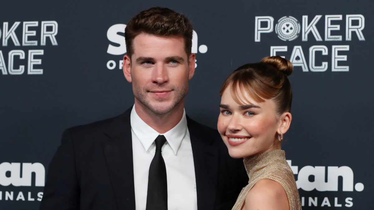 Liam Hemsworth and Gabriella Brooks attends the Australian premiere of Poker Face. Liam is wearing a suit and black tie and Gabriella is wearing a gold halter neck dress and her hair is in a bun.