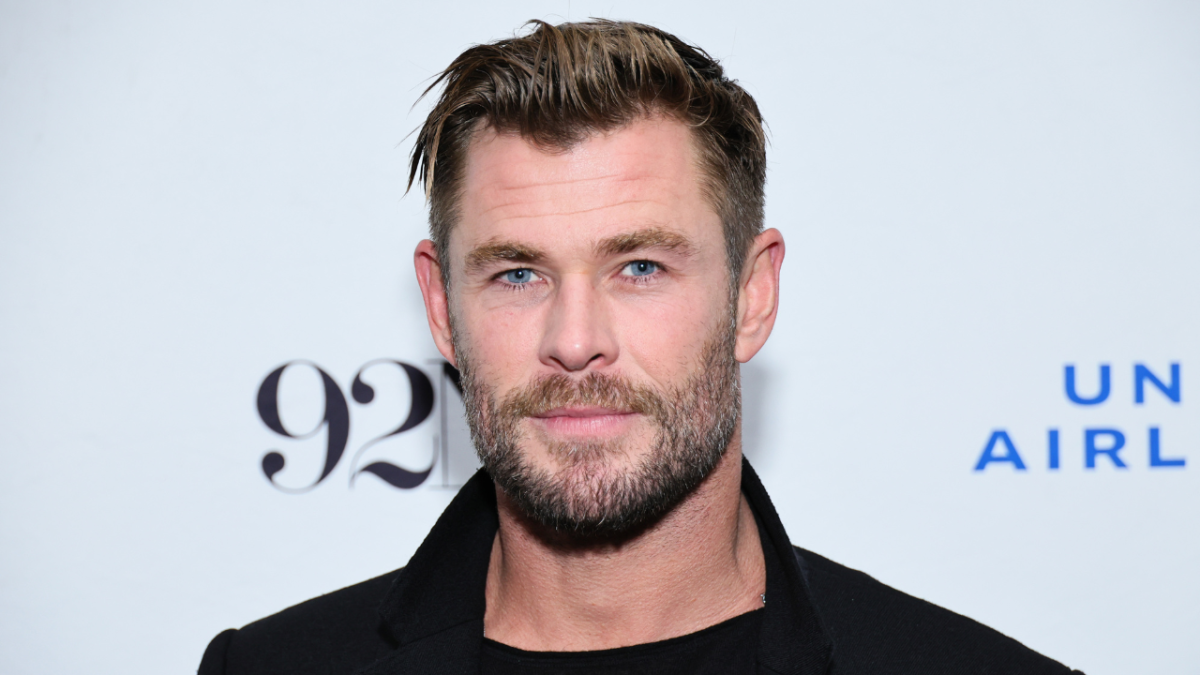 Actor Chris Hemsworth in a black coat and black shirt standing in front of pale blue background.