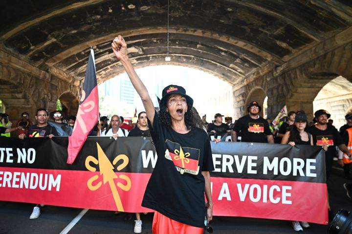 People participate in an Invasion Day protest in Sydney, Australia. A sign reading "We deserve more than a voice" is held by protesters and a woman raises her fist in support.