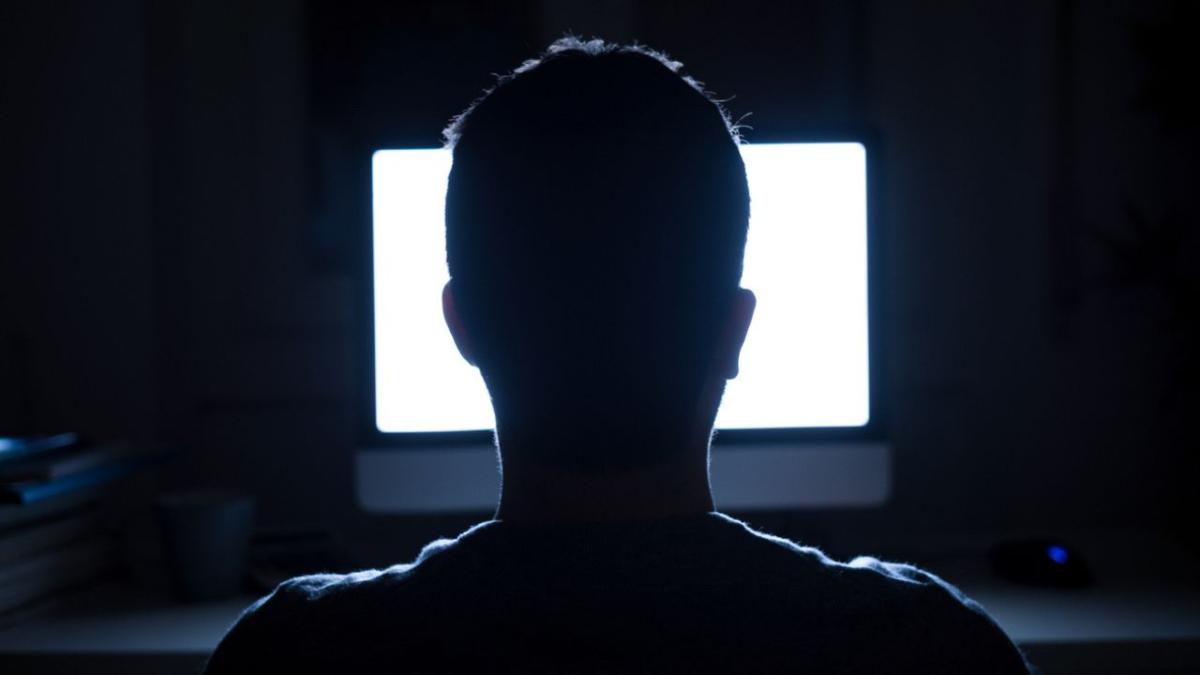 A man sitting at a desk, staring at a bright computer screen with the lights off around him. headline of the story: Why Don't We Treat Non-Consensual Deepfake Porn Of Women As Rape?