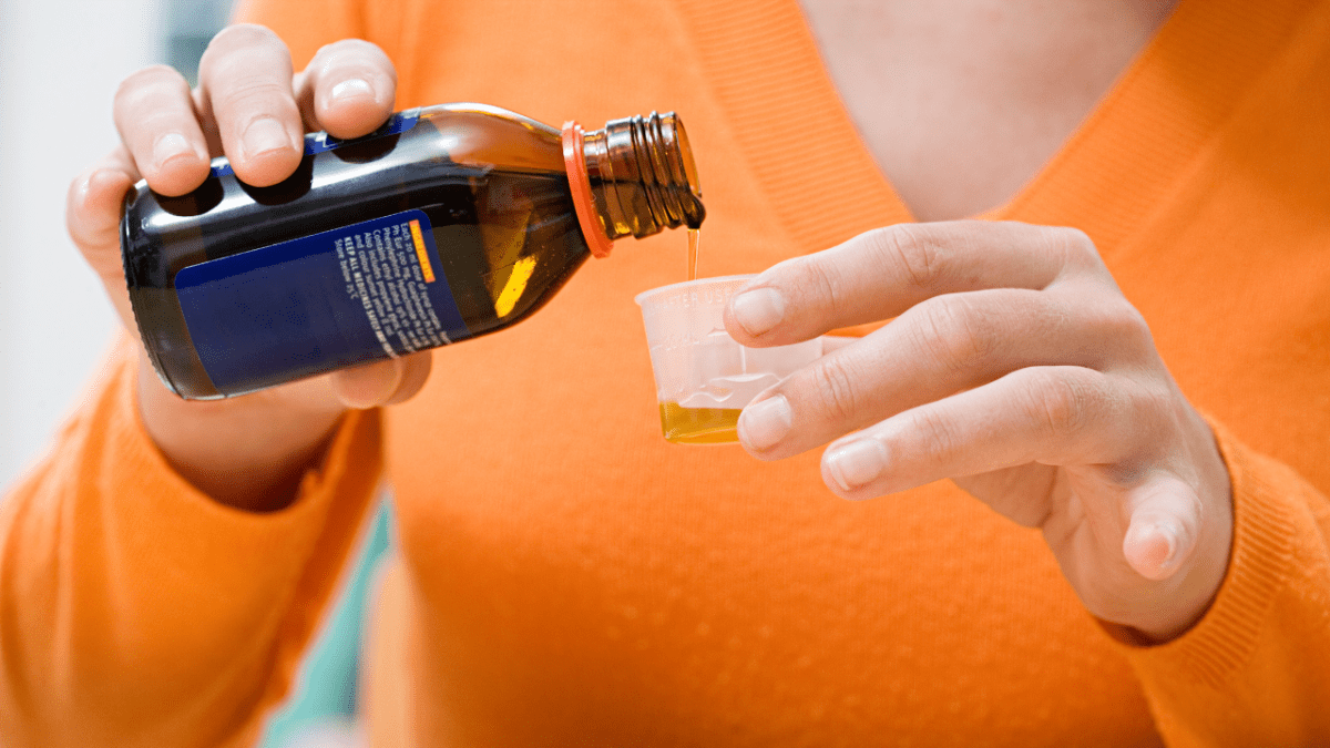 Cough medicine recall: woman wearing an orange jumper pours cough syrup into a measuring cup