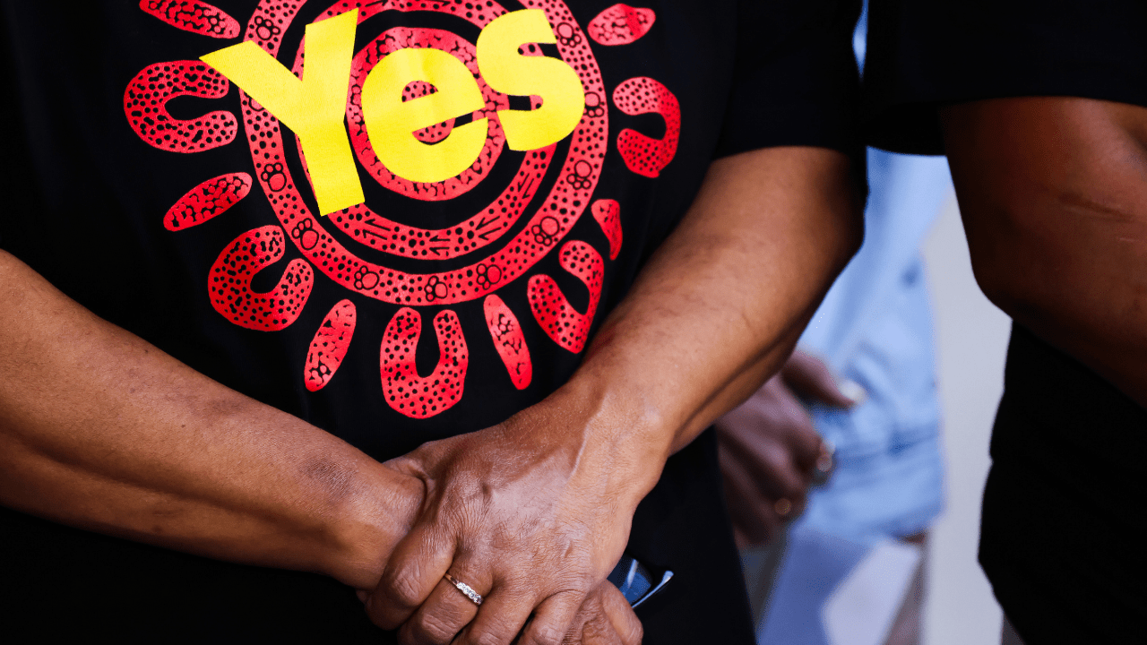 The Yes23 or the Yes Campaign's 'Week Of Silence' Following The Referendum Is Getting Blowback, So We Asked A Psychologist About It. Image below is of a woman's torso. she is wearing a yes campaign shirt and is holding her hands under her chest.