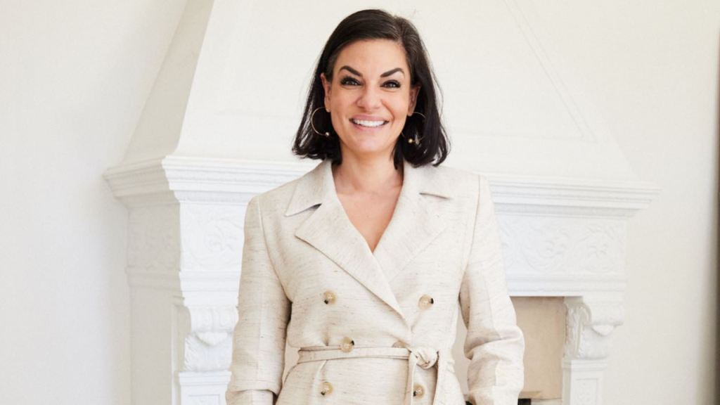 Real Housewives of Sydney star Nicole O'Neil in a cream coat standing in front of a white fireplace.