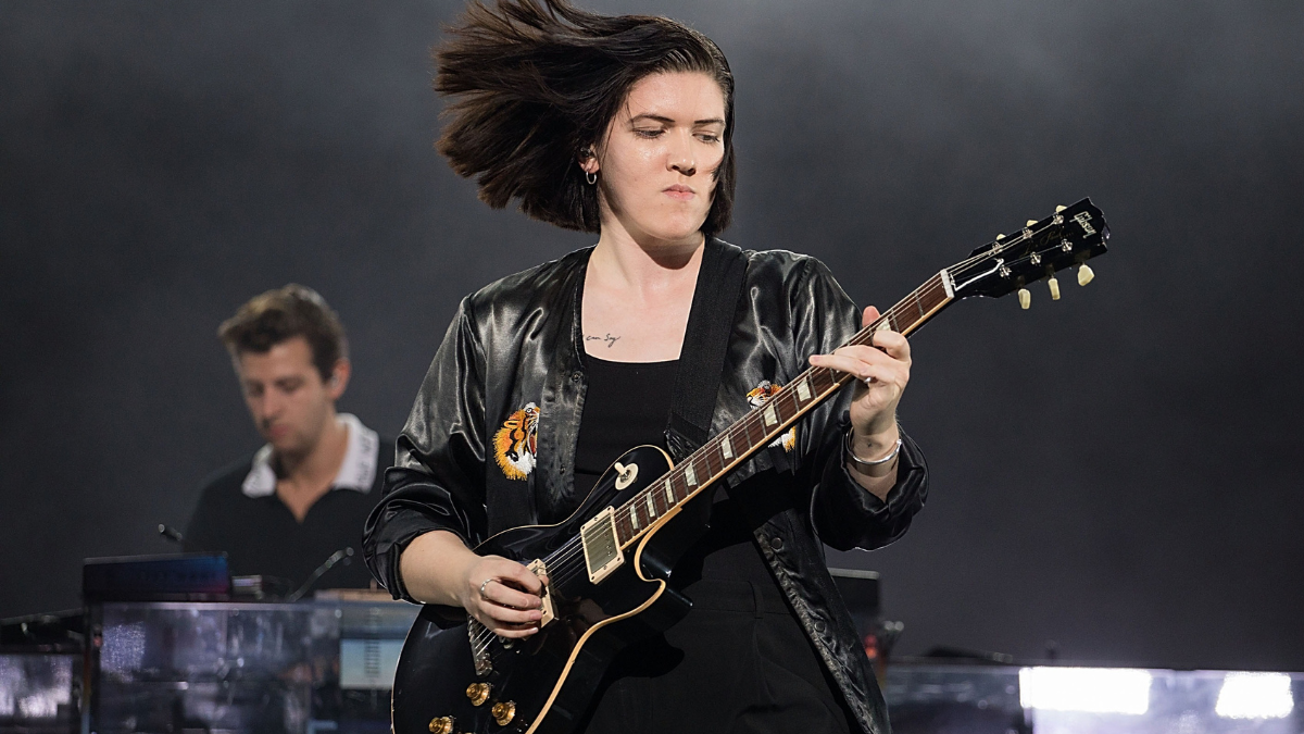 Romy Madley Croft performs onstage with The xx, wearing black clothing and playing guitar